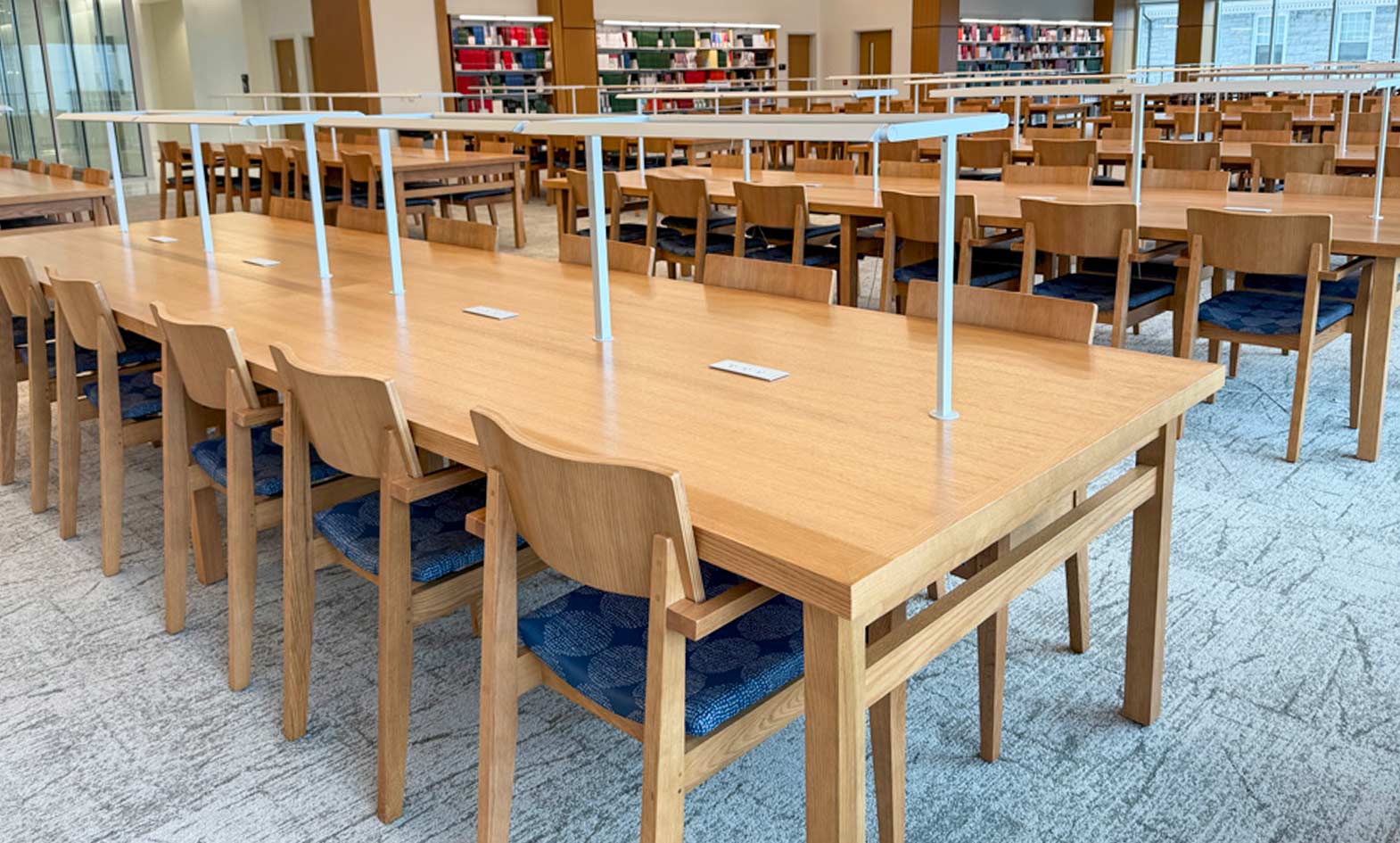 Long durable wooden tables and chairs are arranged in rows in a spacious library study area with bookshelves in the background.